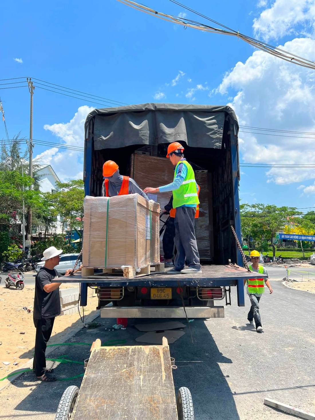 Hai nhân công áo phản quang dỡ kiện pallet bọc màng co từ thùng xe tải